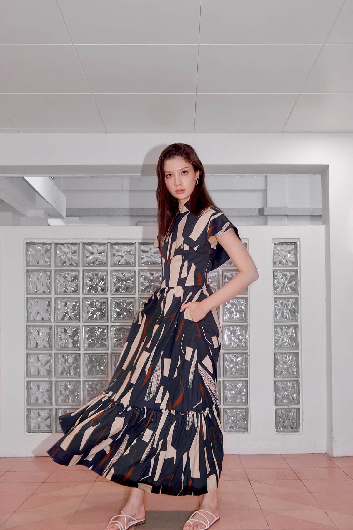 Tall Eurasian model standing in front of glass feature wall, her left hand in the side concealed pocket of her dress. Her dress is long and has a cheongsam collar. It is blue in color with cream rectangles and maroon stripes.
