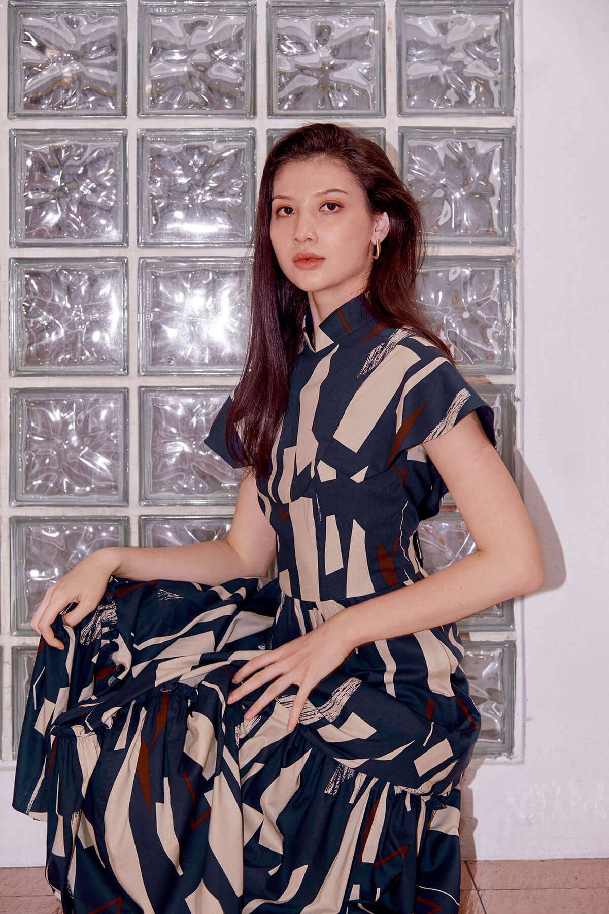 Eurasian model squats against a wall. She is wearing a long cheongsam dress, with her right arm resting on her right knee and left arm on her left thigh. The qipao dress has cape like sleeves and a gathered skirt in maxi length.