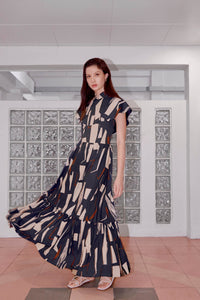 Tall Eurasian model standing in front of glass feature wall, her left hand by her side. Her dress is long and has a cheongsam collar. It is blue in color with cream rectangles and maroon stripes.