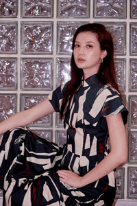 Eurasian model squats against a wall. She is wearing a long cheongsam dress, with her right arm resting on her right knee, which is slightly elevated. Her left arm is rested on her left thigh. The qipao dress is blue and has a mandarin collar.
