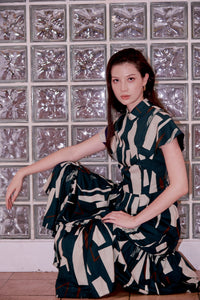 Eurasian model squats by a wall, posing for a picture. She is wearing a long cheongsam dress, with her right arm resting on her right knee, which is slightly elevated. The dress is full length and had a standup mandarin collar.