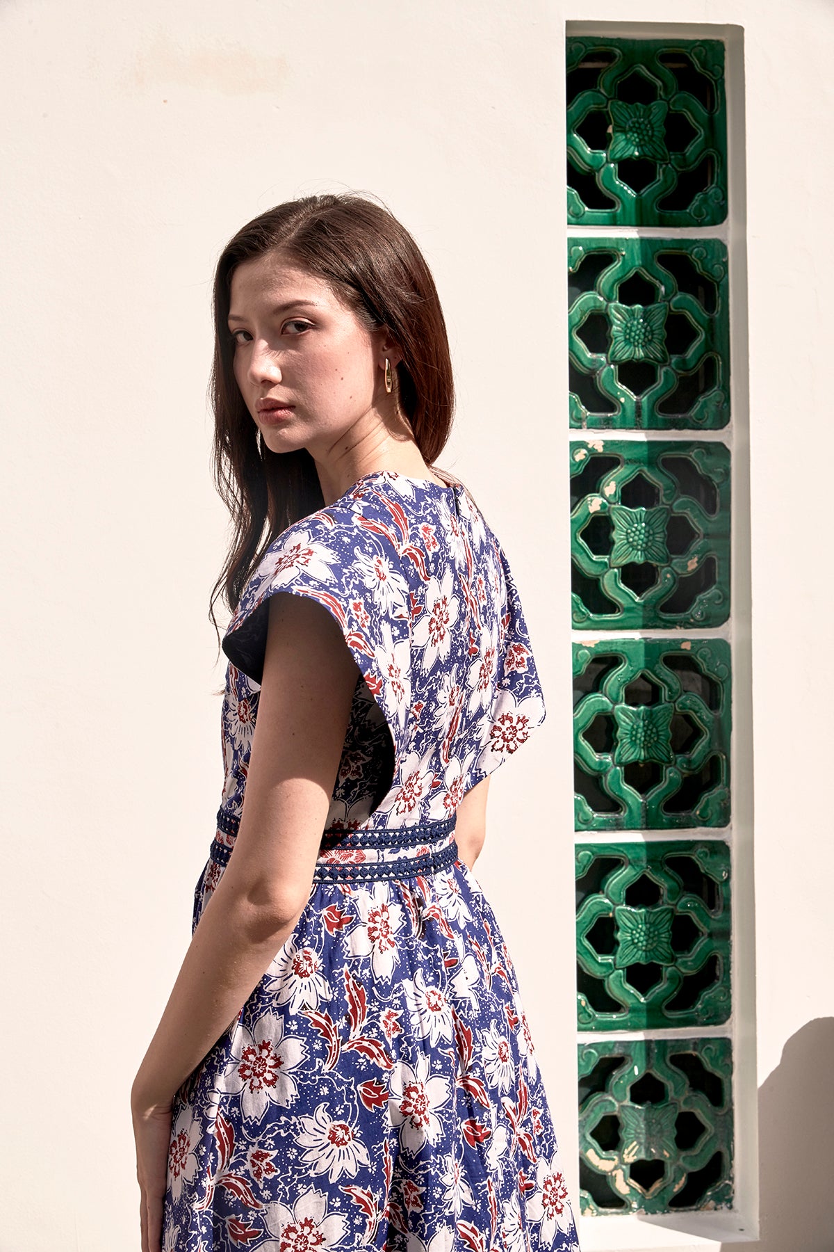 Eurasian female model, modelling a blue and red batik dress and lace trimmed waist. Picture showed the back view of the dress and with her head looking back at the camera. Dress has white flowers against blue background.