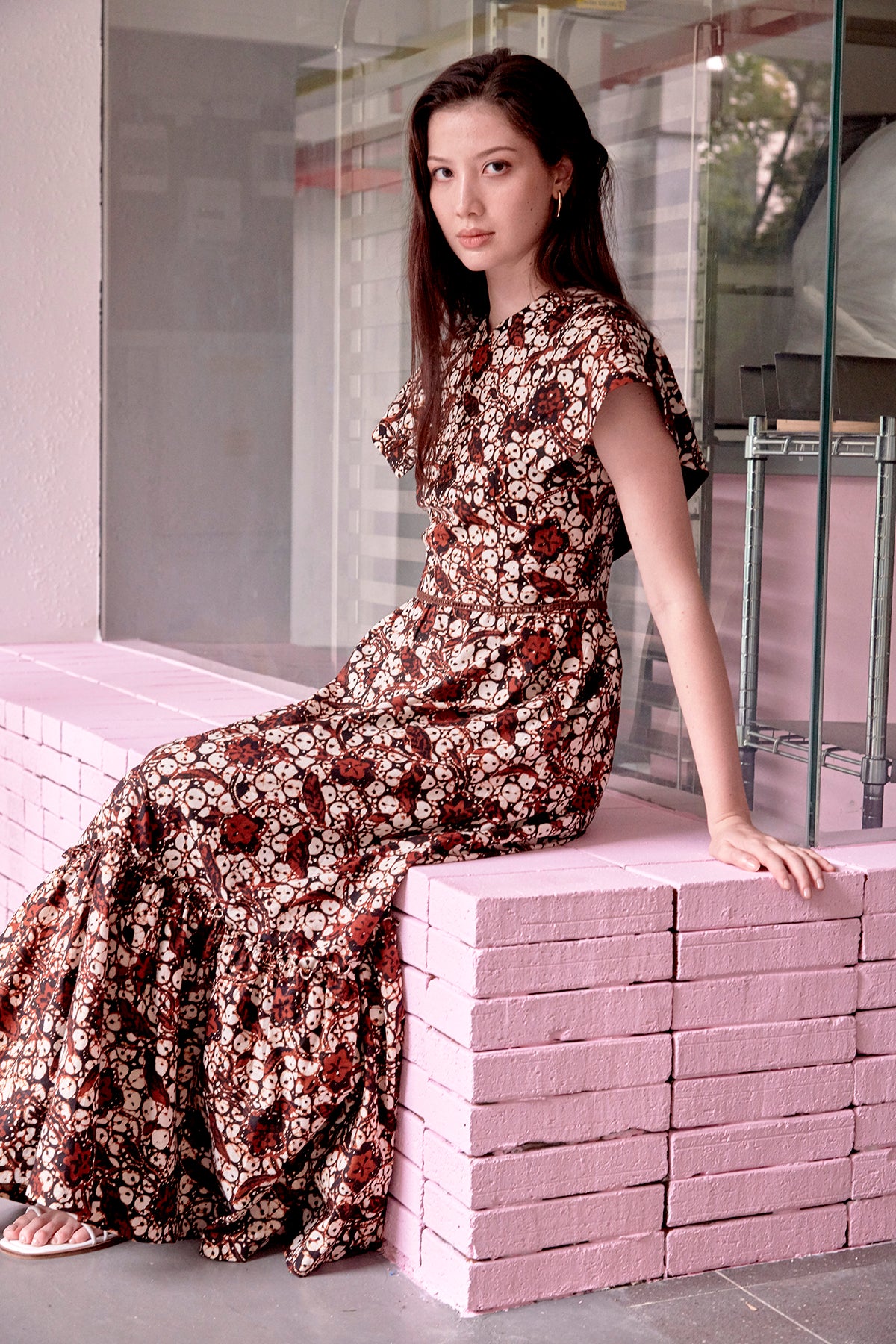 Eurasian model with long hair,  seated on a pink ledge, wearing brown batik full length dress with cape sleeves