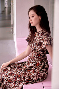 Side view of eurasian model with long hair, seated on a pink ledge, wearing maxi length brown batik dress with cape sleeves. Dress has bohemain motifs