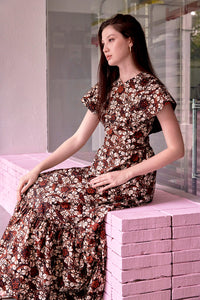 Side view of long haired eurasian model, seated on a pink ledge, wearing maxi length brown batik dress with cape sleeves. Fabric of dress is chocolate brown and white batik with bohemain motifs