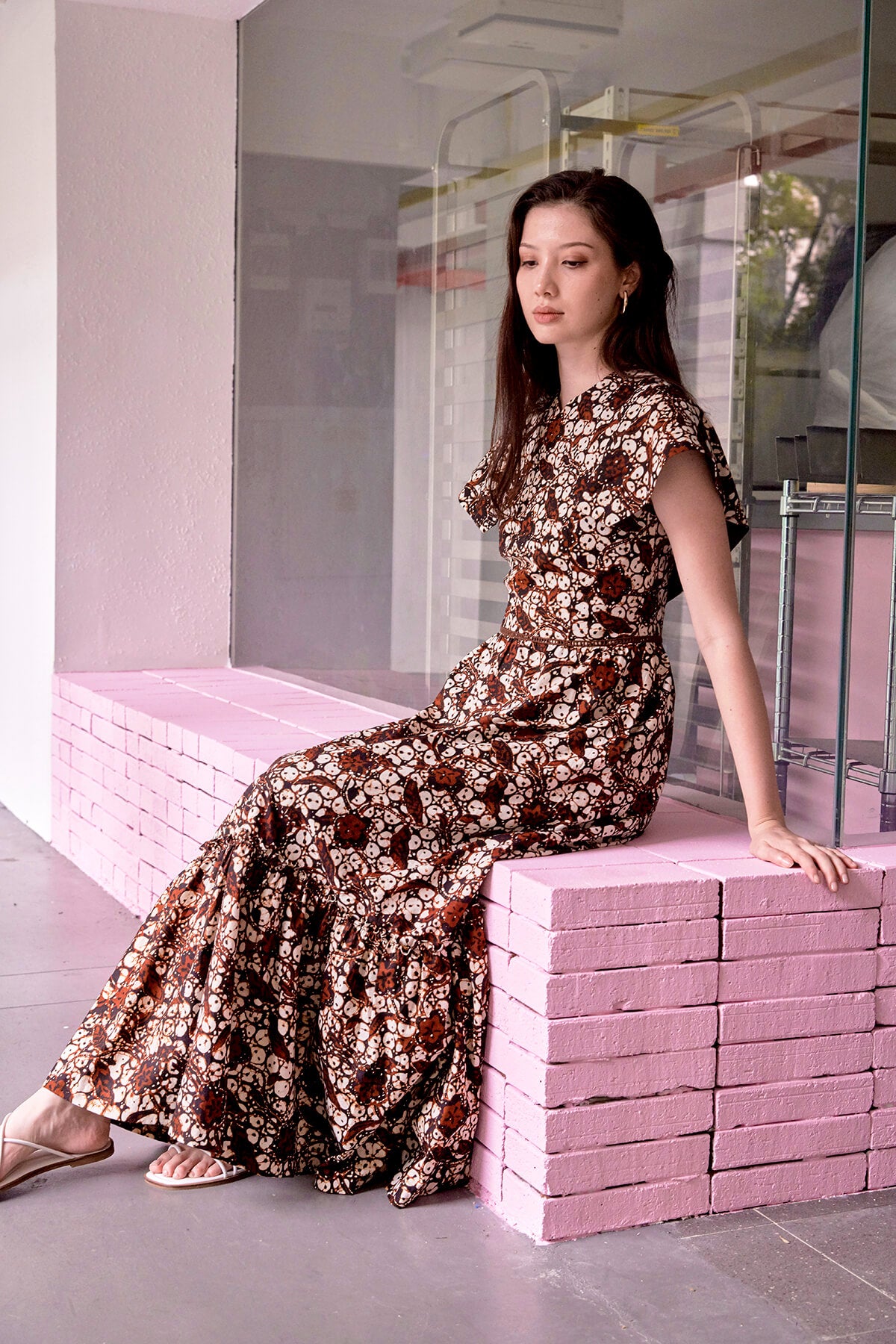Eurasian lady sitting on a pink ledge at Everton Park Singapore. She wears a maxi batik dress by Singapore designer brand Dayglow. She is also wearing a pair of gold earrings and white strappy sandals, and has her gaze on the floor.