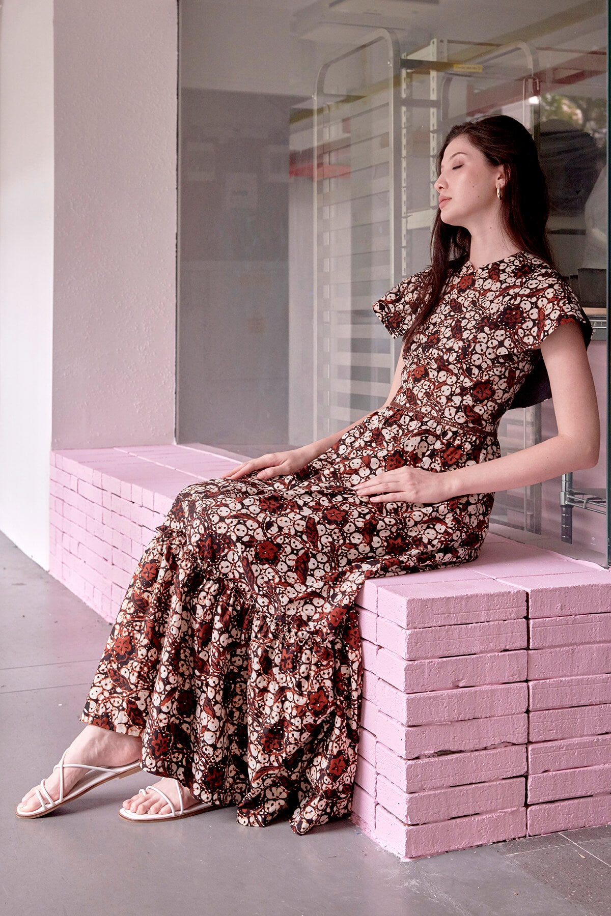 Side view of long haired eurasian model, seated on a pink ledge, wearing maxi length brown batik dress with cape sleeves. Fabric of dress is wax resist cotton also known as batik