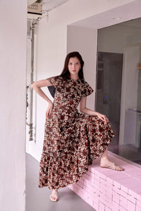 Tall Eurasian model, with left leg on a pink ledge, standing with right hand on her waist. She is wearing a long batik dress, with brow ansd white wax resist prints and wearing white strappy sandals.