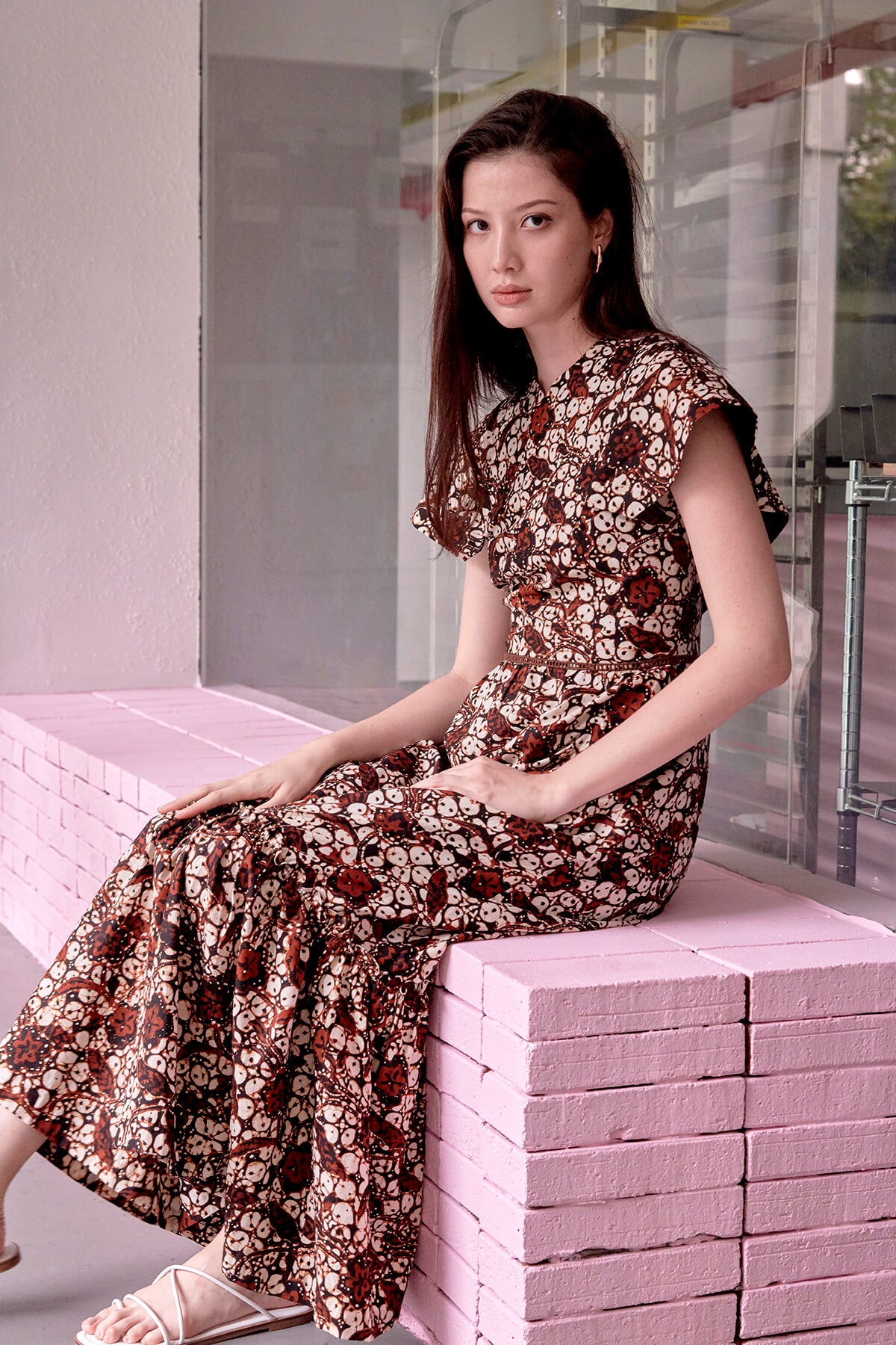 Young eurasian female lady, seated on pink ledge, modelling a long batik dress with brown and white batik prints.