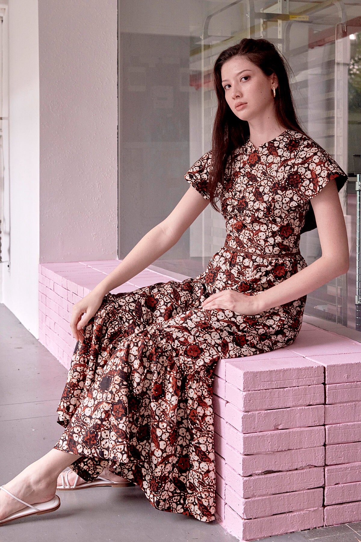 Singaporean female model seated in a brown batik long dress, looking into camera with long hair and gold earrings