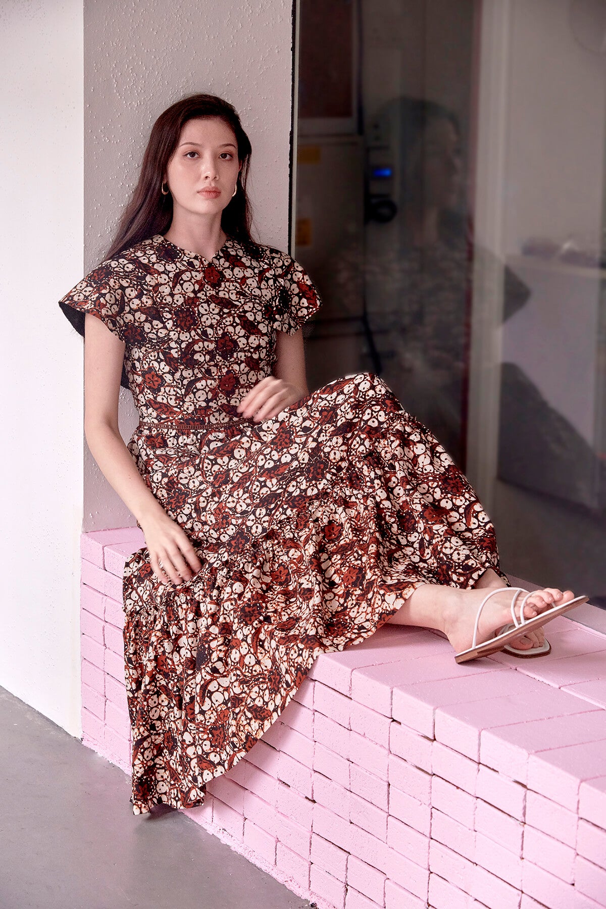 Eursian woman sitting on a pink ledge, legs stretched out and looking relax in a long brown batik dress. Dress has sleeves like a cap and a long tiered skirt.