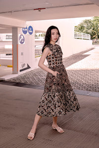 Chinese model posing with her side view facing the camera. She is wearing a brown batik dress, in mid calf length and flared skirt. She has her right hand in the side concealed pocket of the dress. Dress has four different pieces of batik fabric.