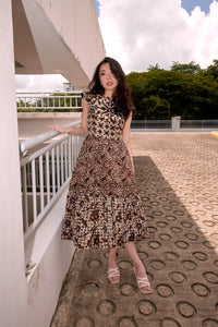 Chinese lady with long wavy hair, standing beside railing, wearing a midi length fit and flare batik dress. Dress has sleeves, gathered skirt with gathered tier at the hemline. Batik is hand stamped and is popular in Indonesia and Singapore.