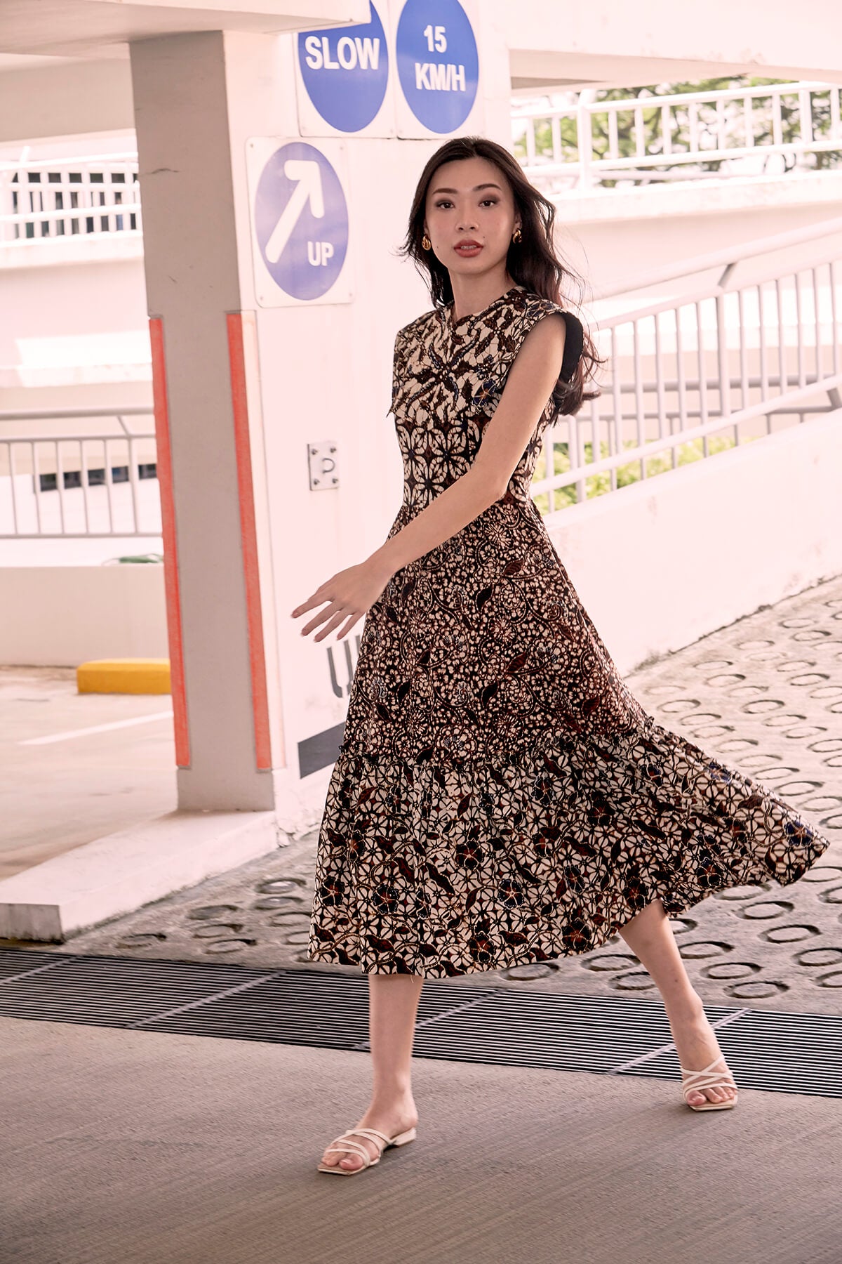 Chinese model taking a long stride at a carpark. She is posing for a picture wearing a brown batik dress, in mid calf length and flared skirt. She has her left arm in front of her. She has long wavy hair.
