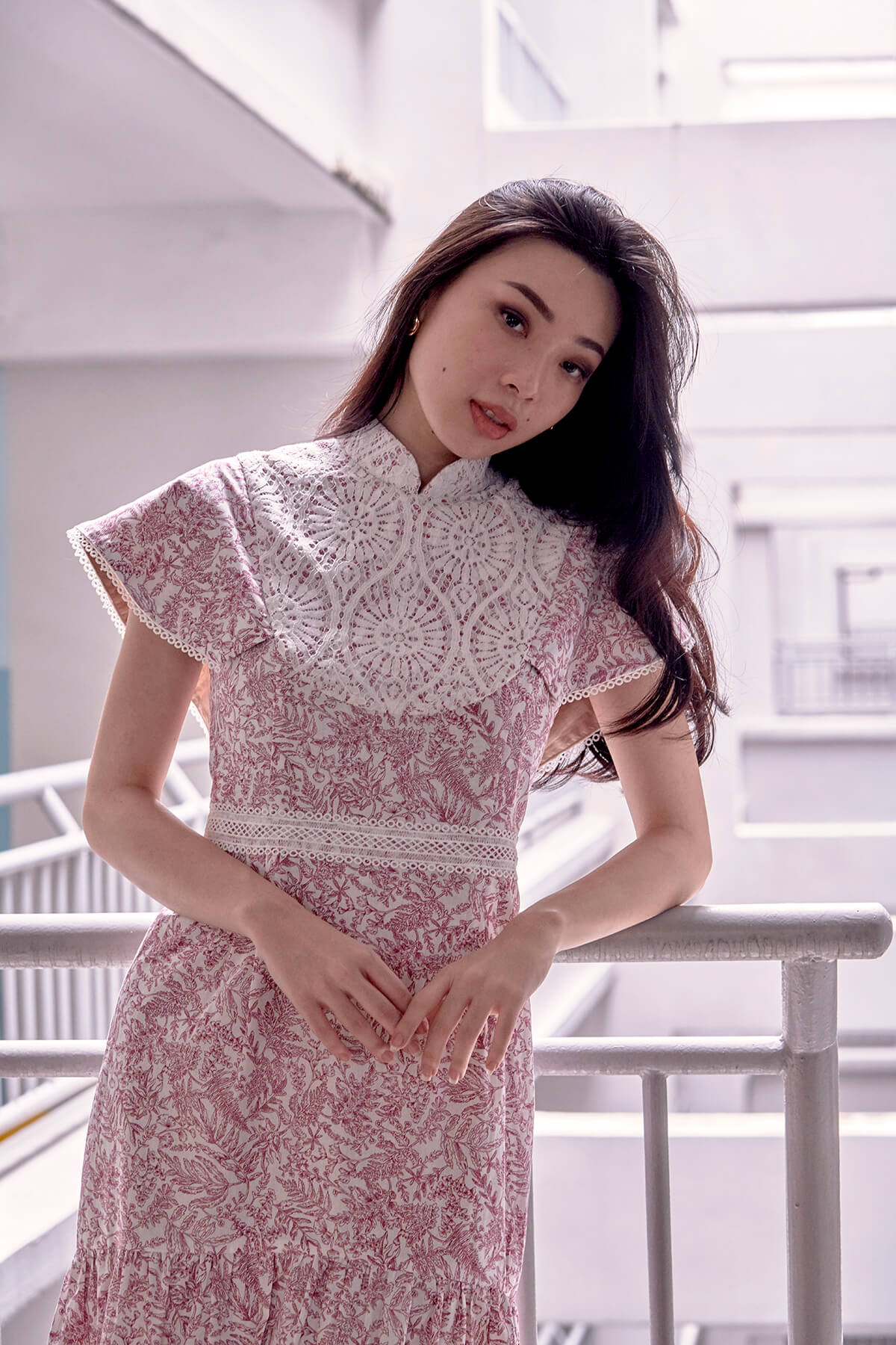 Chinese lady wearing a red and white foliage print qipao dress. Dress has cape like sleeves, lace overlay details and stand up mandarin collar. She tilts her head and has her elbow on the railings.