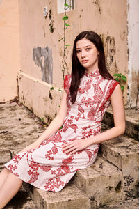 Eurasian lady seated on a stair, with her left elbow resting on the stair. She wears a red and white batik cap cheongsam dress, with long brown hair. The dress has red leaves and flowers printed using wax resist method, A-line shin length skirt and panel as sleeves.  