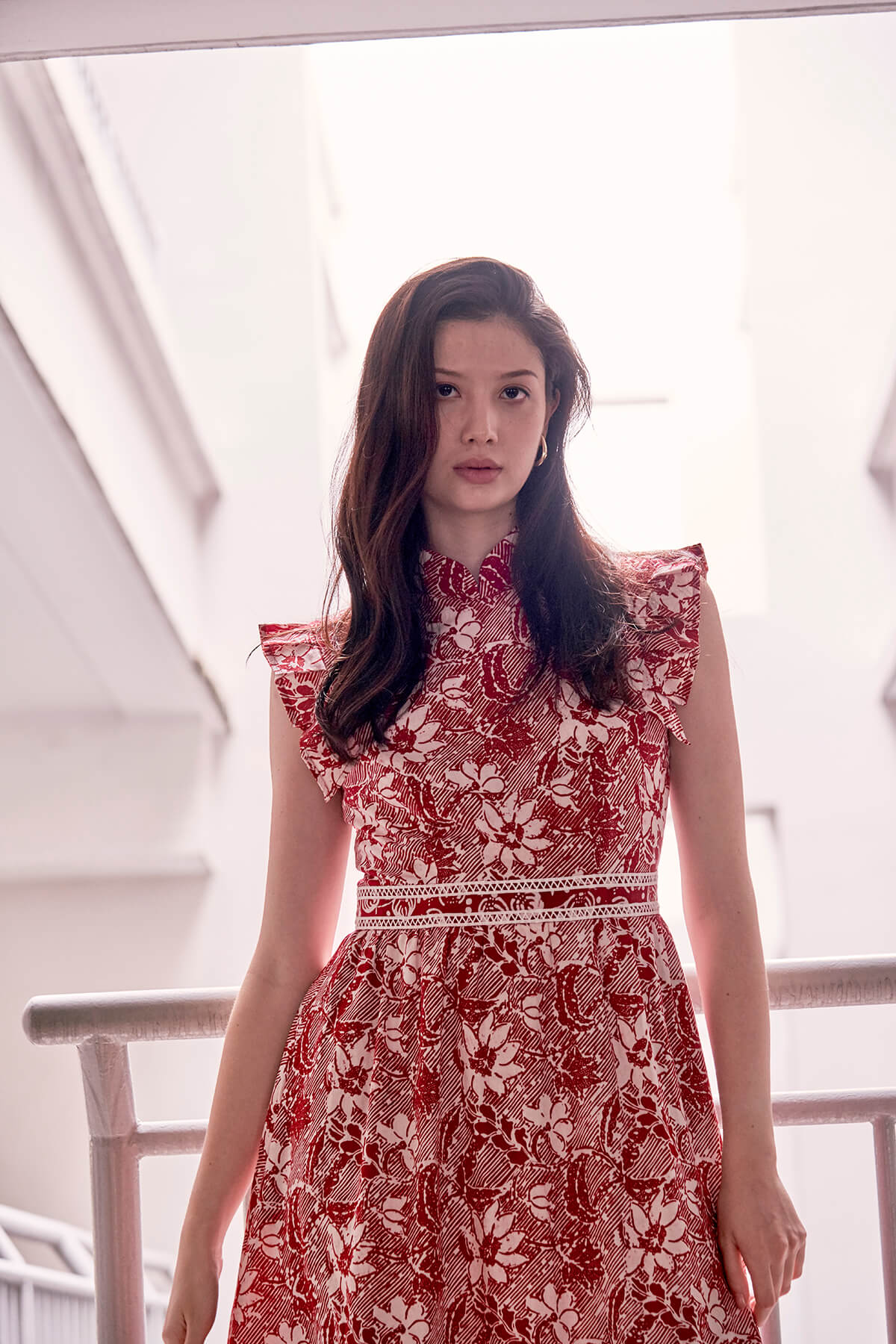 Half body view of young Eurasian  model, with gold earrings and red white batik dress. Dress has a stand up mandarin collar, and pleated trimming which looked like sleeves. There is also a waistband with lace trimming.